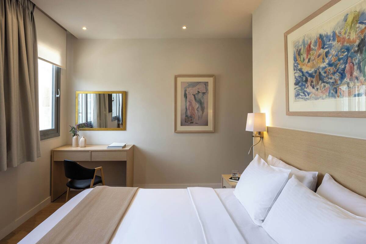 Ein Hotelzimmer mit Bett, Schreibtisch, Stuhl, Lampe, einem Gemälde und einem Fenster mit Vorhängen.