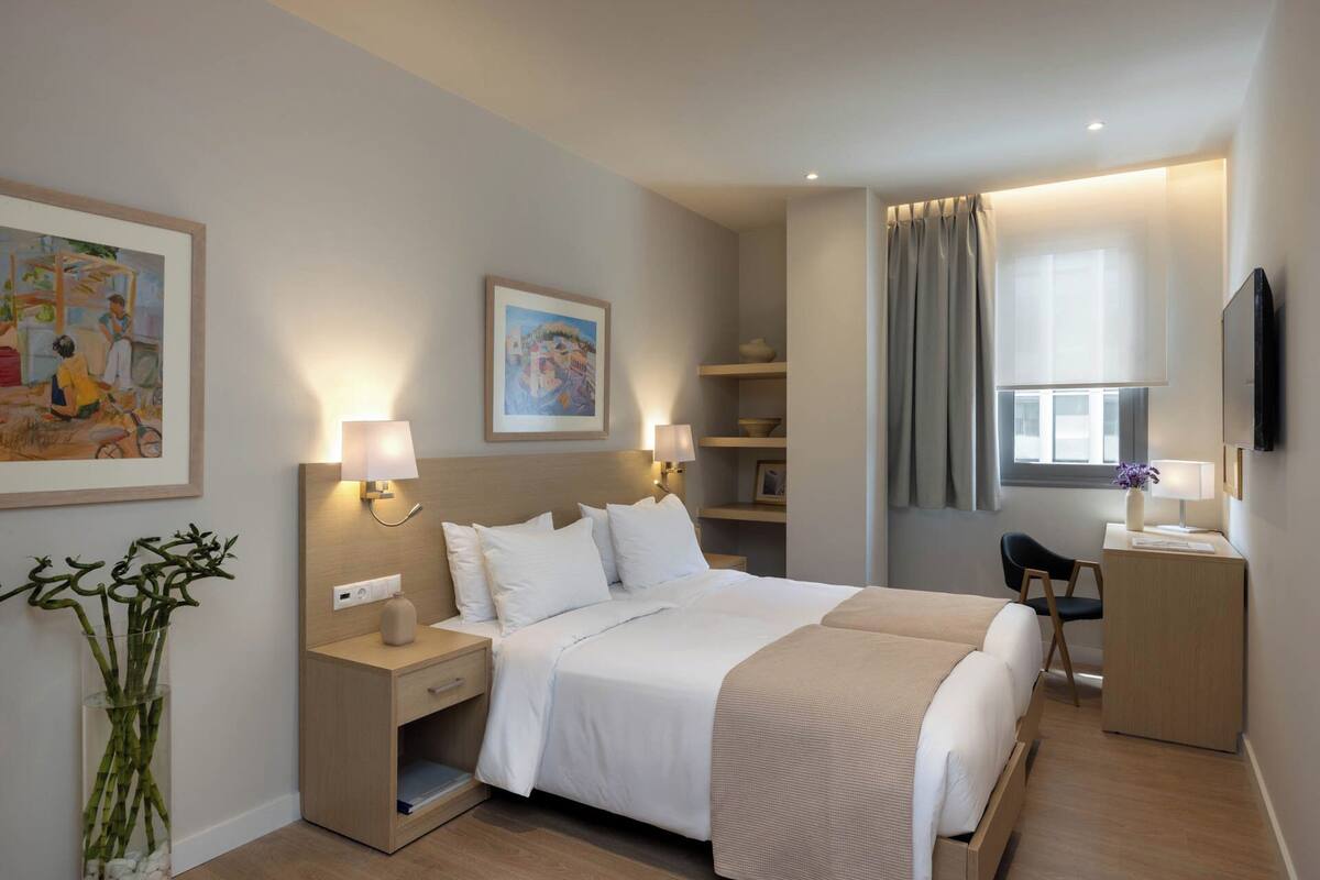 Ein modernes Hotelzimmer mit einem großen Bett, Nachttischen, einem Schreibtisch und einem Fernseher. An den Wänden befinden sich Gemälde und es gibt ein Fenster mit Vorhängen.