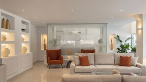 Una sala de estar moderna con un sofá, sillones y una mesa de centro. Hay jarrones decorativos en estanterías y una planta a la derecha.