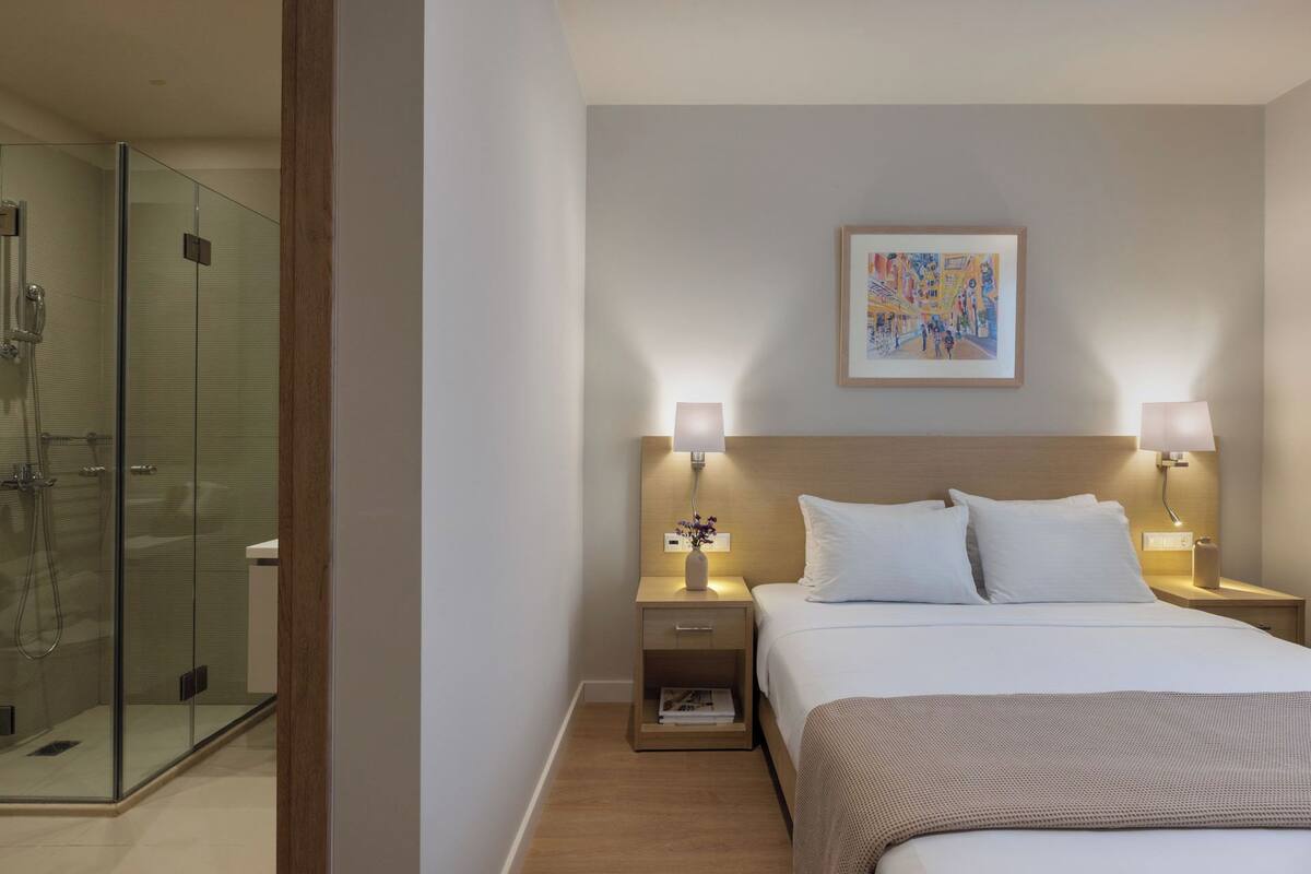 Ein modernes Hotelzimmer mit einer gläsernen Duschkabine, einem Bett mit weißen Bettwäsche, zwei Nachttischlampen und einem gerahmten abstrakten Gemälde an der Wand.