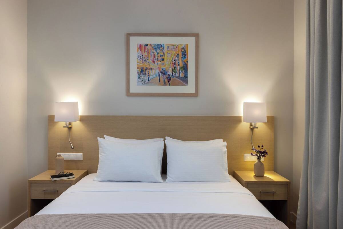 Ein Hotelzimmer mit einem großen Bett, zwei Nachttischlampen, einem gerahmten abstrakten Gemälde und einem Nachttisch mit einer Blumenvase.