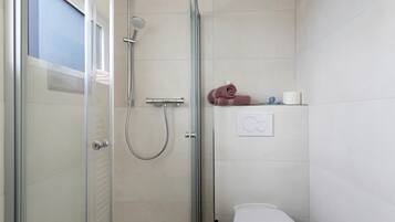 A modern bathroom with a glass shower enclosure, a toilet, and a shelf with towels and toiletries.