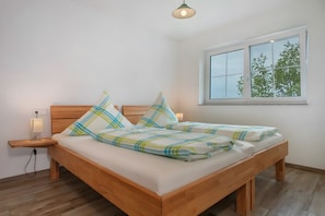 Un lit double recouvert d’une couverture à carreaux, une table de chevet avec une lampe et une fenêtre donnant sur les arbres.