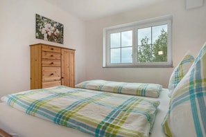 Une chambre à coucher avec un lit, deux oreillers, une commode en bois, une fenêtre et un tableau représentant des motifs floraux accroché au mur.