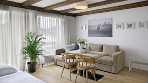Ett modernt vardagsrum med en soffa, två trästolar, ett matbord, en krukväxt och ett stort fönster med tunna, genomskinliga gardiner.