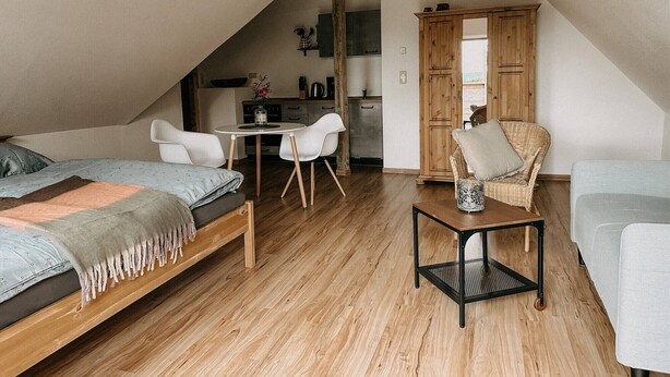 Een slaapkamer in loftstijl met een schuine kap, een bed, een klein tafeltje, een stoel en een kitchenette op de achtergrond.