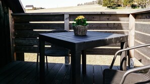 Een houten terras buiten met een tafel en stoelen, een rieten mand met bloemen en uitzicht op bomen en open land.