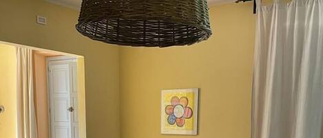 A room with a bed, two chairs, a small table, a hanging light fixture, and a door leading to another room.