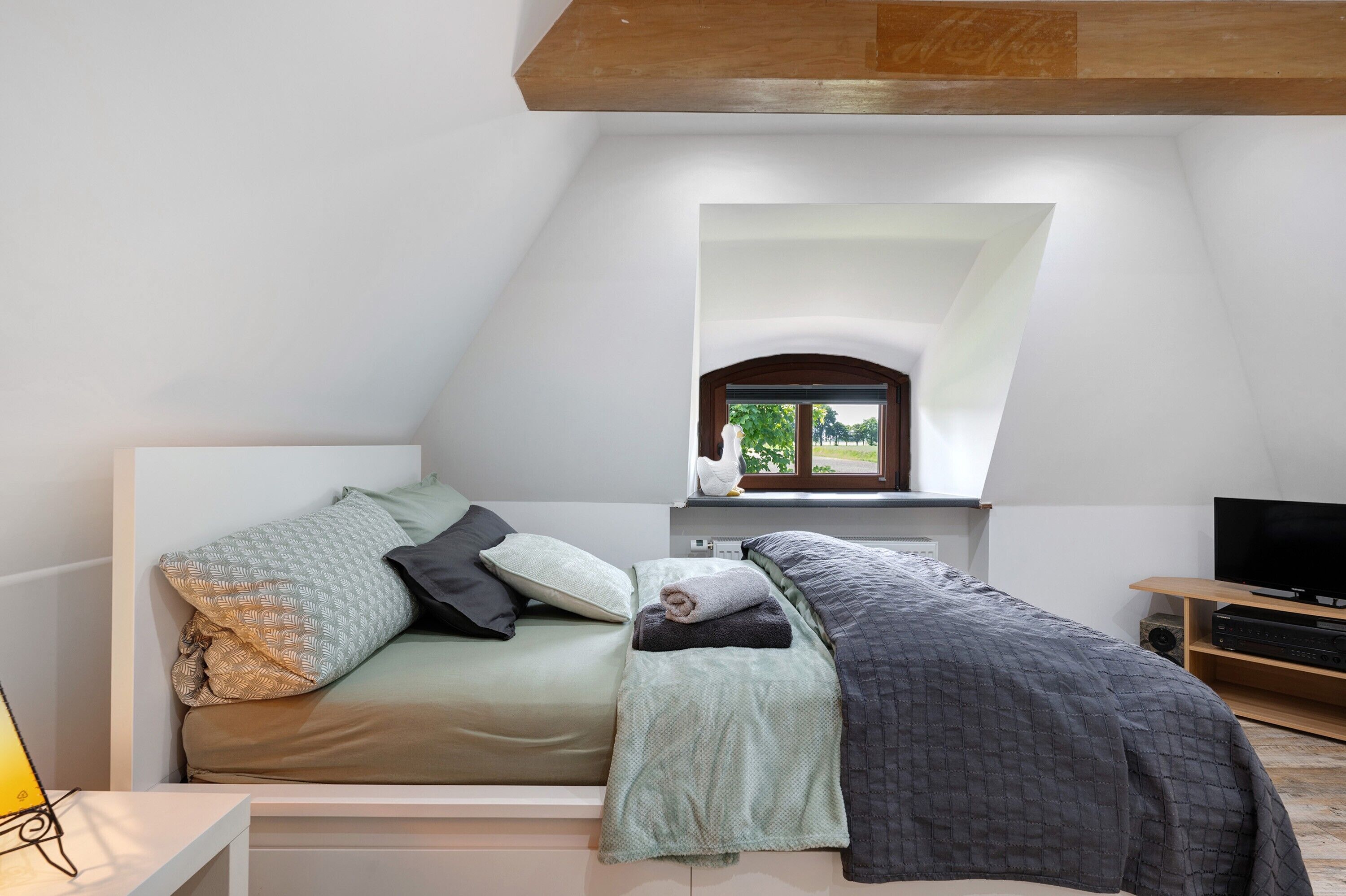A bedroom with a bed, pillows, a blanket, a TV, and a window with a view.