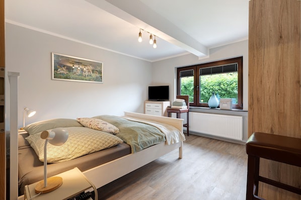 Ein Schlafzimmer mit Bett, Nachttischlampe, Fernseher, Kommode, Fenster und Heizkörper.