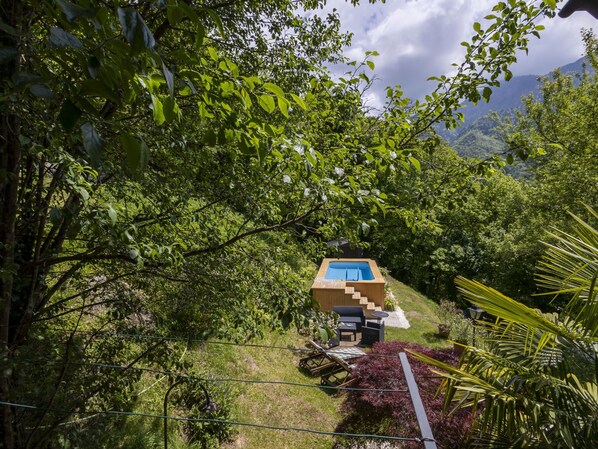 A wooden hot tub with a blue pool, surrounded by lush greenery and a mountain backdrop.