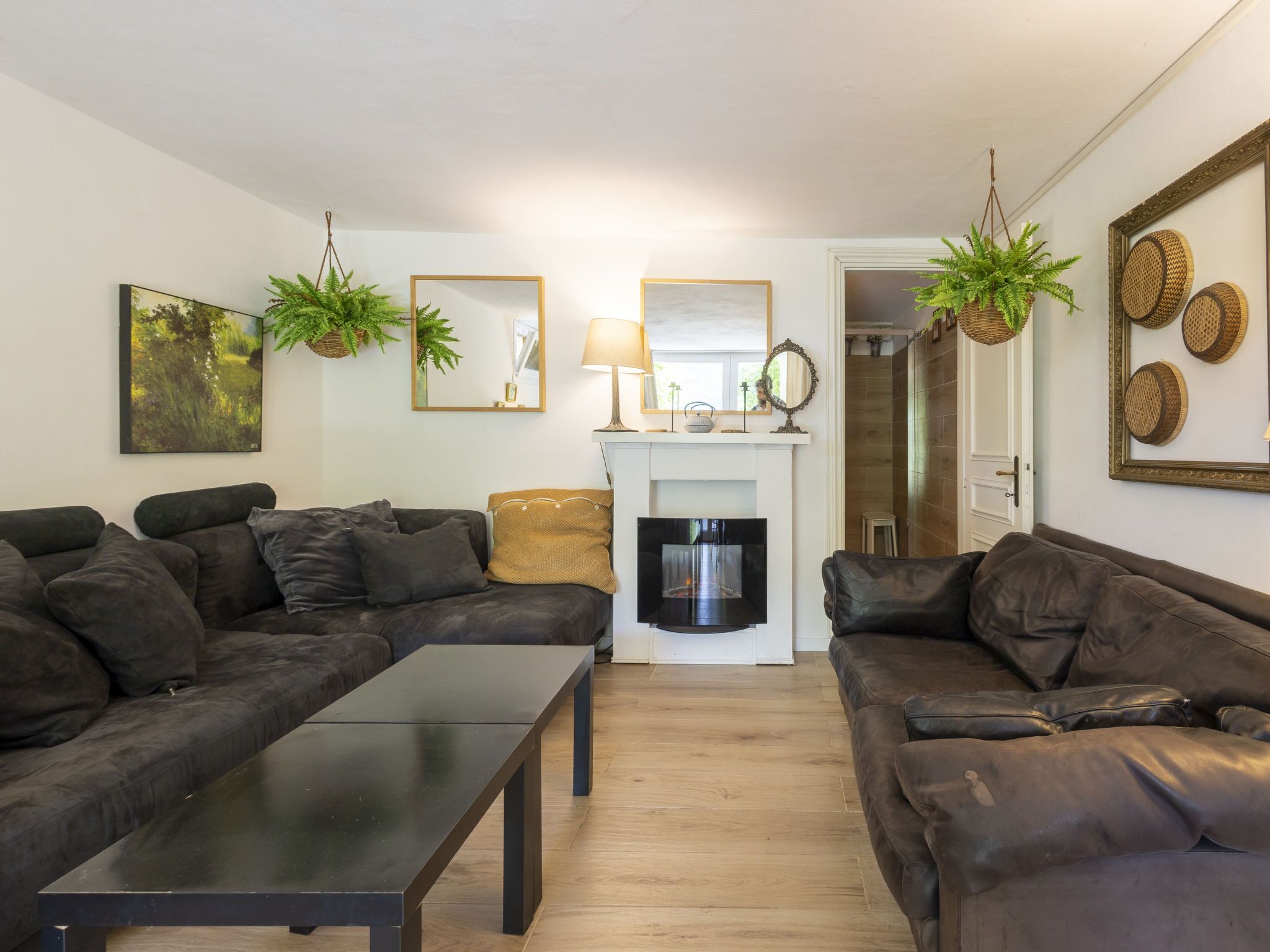 A living room with two dark leather sofas, a black coffee table, and a fireplace. There are framed pictures and plants on the walls.