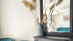 Un fauteuil recouvert d’un tissu de couleur bleu-vert, une table en bois foncé ornée d’un vase contenant des herbes des pampas séchées, et un miroir qui reflète la pièce.