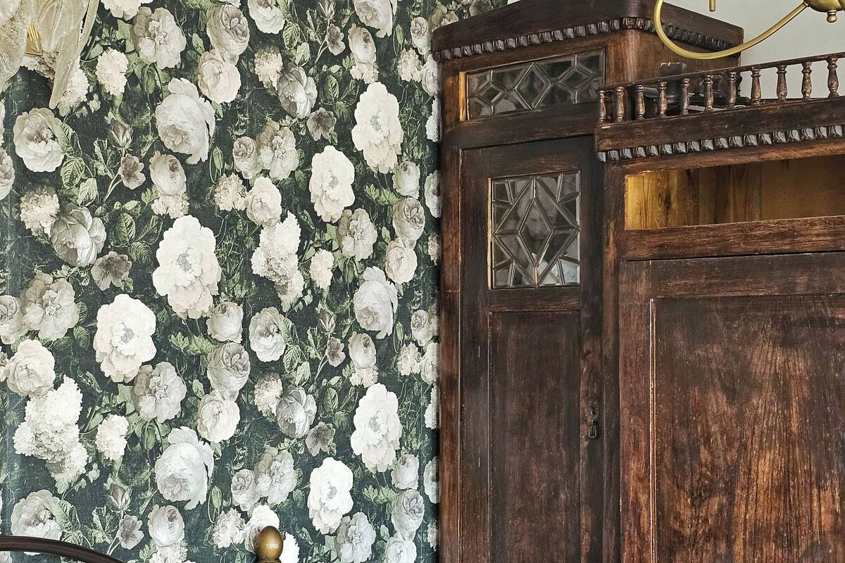 A bedroom with a floral wallpaper, a wooden wardrobe, a bed with a floral bedspread, and a vase with flowers.