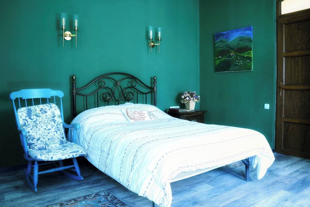 A bedroom with a bed, a rocking chair, a nightstand with a vase, and a painting on the wall.