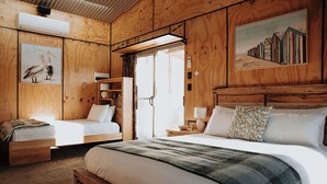 Une chambre de style cabane en bois, avec deux lits, une grande fenêtre et un tableau représentant des cabanes de plage.