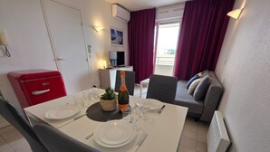 Un coin repas avec une table blanche dressée pour deux personnes, un appareil électroménager rouge, une plante et une bouteille de champagne.