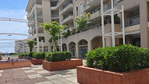 Un immeuble résidentiel à plusieurs étages, doté de balcons, d’une allée pavée et de jardinières rectangulaires agrémentées de verdure.