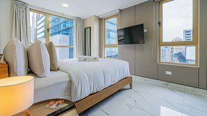 A modern hotel room with a bed, a TV, and large windows offering city views.