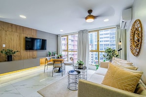 Una sala de estar moderna con un televisor de pantalla plana, un sofá, una mesa de comedor con sillas y ventanas amplias que ofrecen vistas a la ciudad.