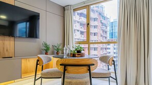 A modern living room with a round dining table, two chairs, a TV mounted on the wall, and a view of high-rise buildings through large windows.
