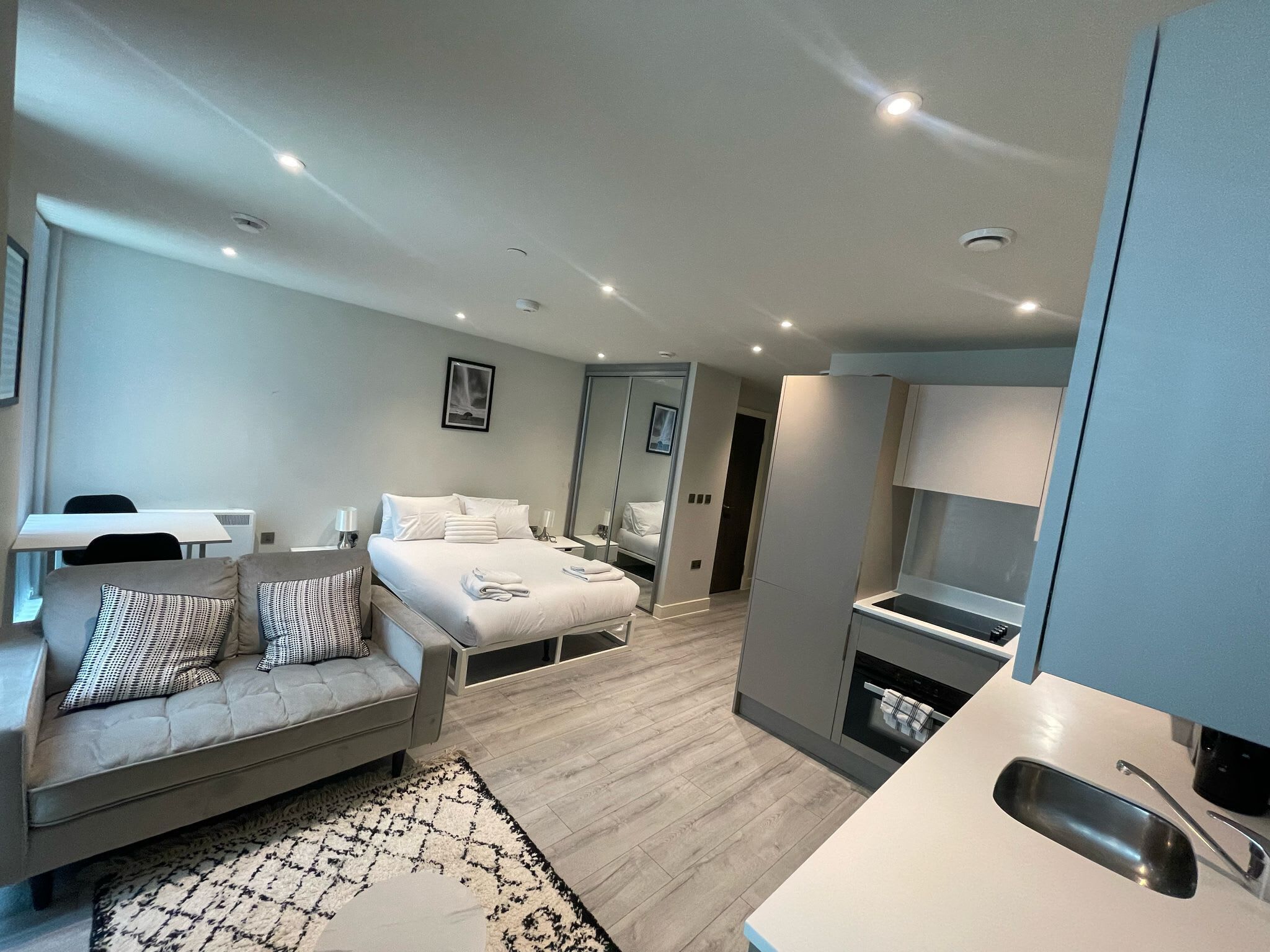 A modern hotel room with a bed, a sofa, a dining table, and a kitchenette.