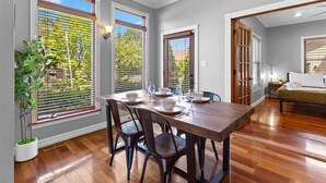 Comedor con mesa de madera dispuesta para cuatro, flanqueado por ventanas amplias con persianas, una planta en maceta y vista al verdor exterior.