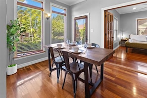 A dining area with a wooden table set for four, flanked by large windows with blinds, a potted plant, and a view of greenery outside.