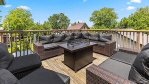 Una terraza al aire libre con un hogar a leña integrado, muebles de mimbre y vista a una casa en el fondo.