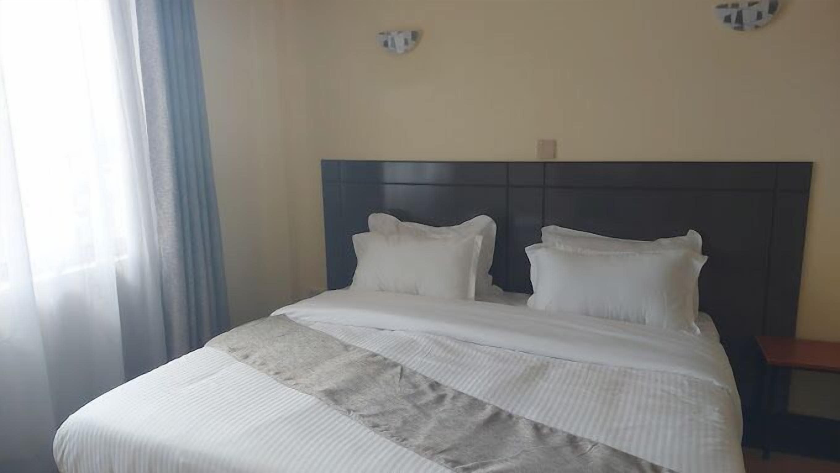 A neatly made bed with a headboard, a window with curtains, and a wall-mounted light fixture.