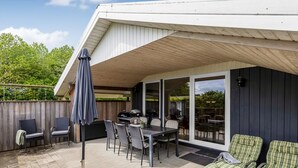 Eine überdachte Terrasse mit Tisch, Stühlen, einem Grill und Liegestühlen.