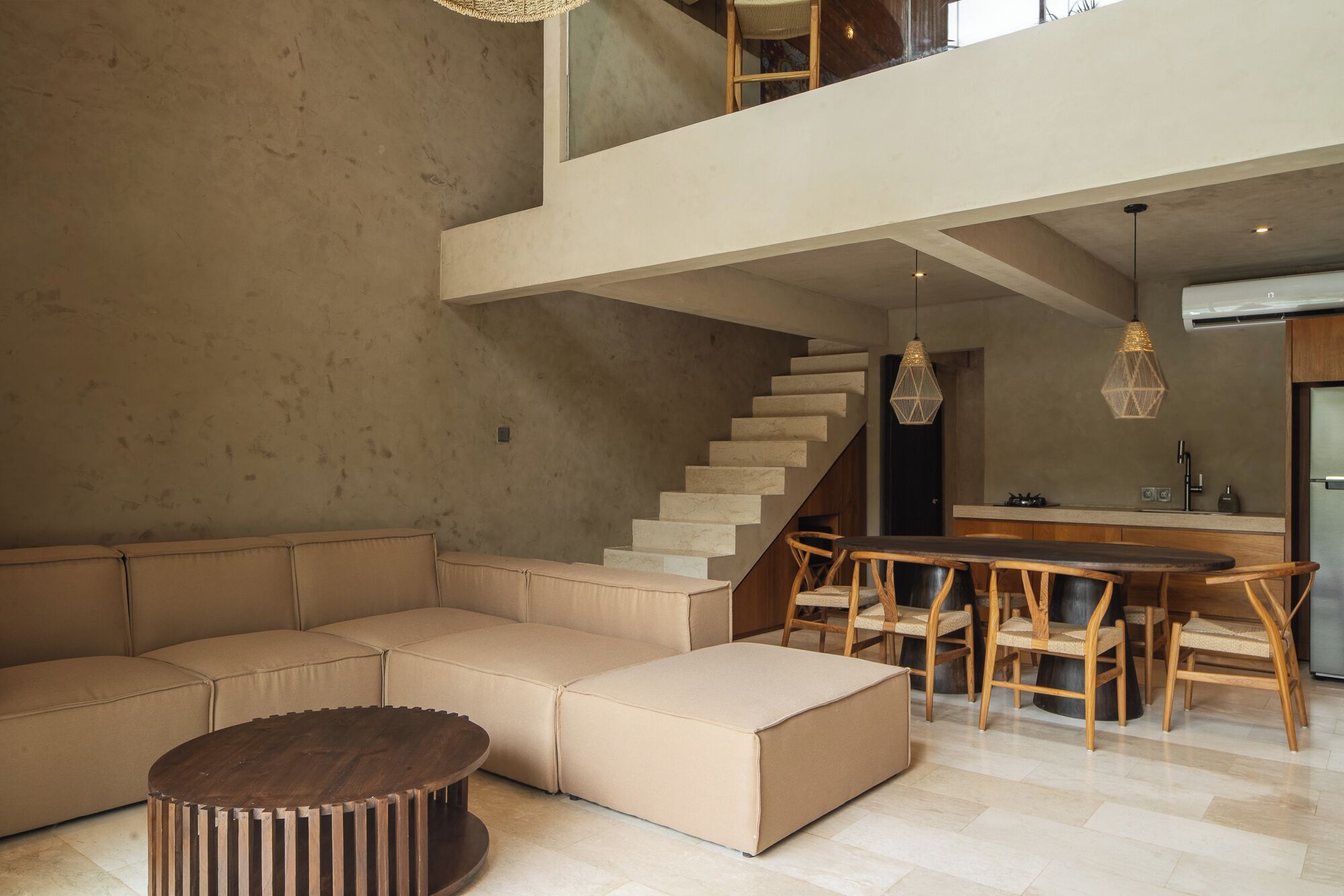 Een moderne woonruimte met een modulaire bank, een ronde salontafel, een eethoek met houten stoelen, een trap en een keukenhoek.