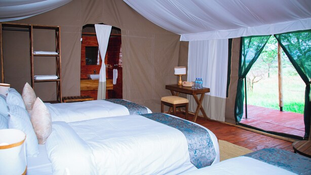 A tent-style room with two beds, a small table with a lamp, and a view of greenery outside. - Mlimwa Camp (Arusha)