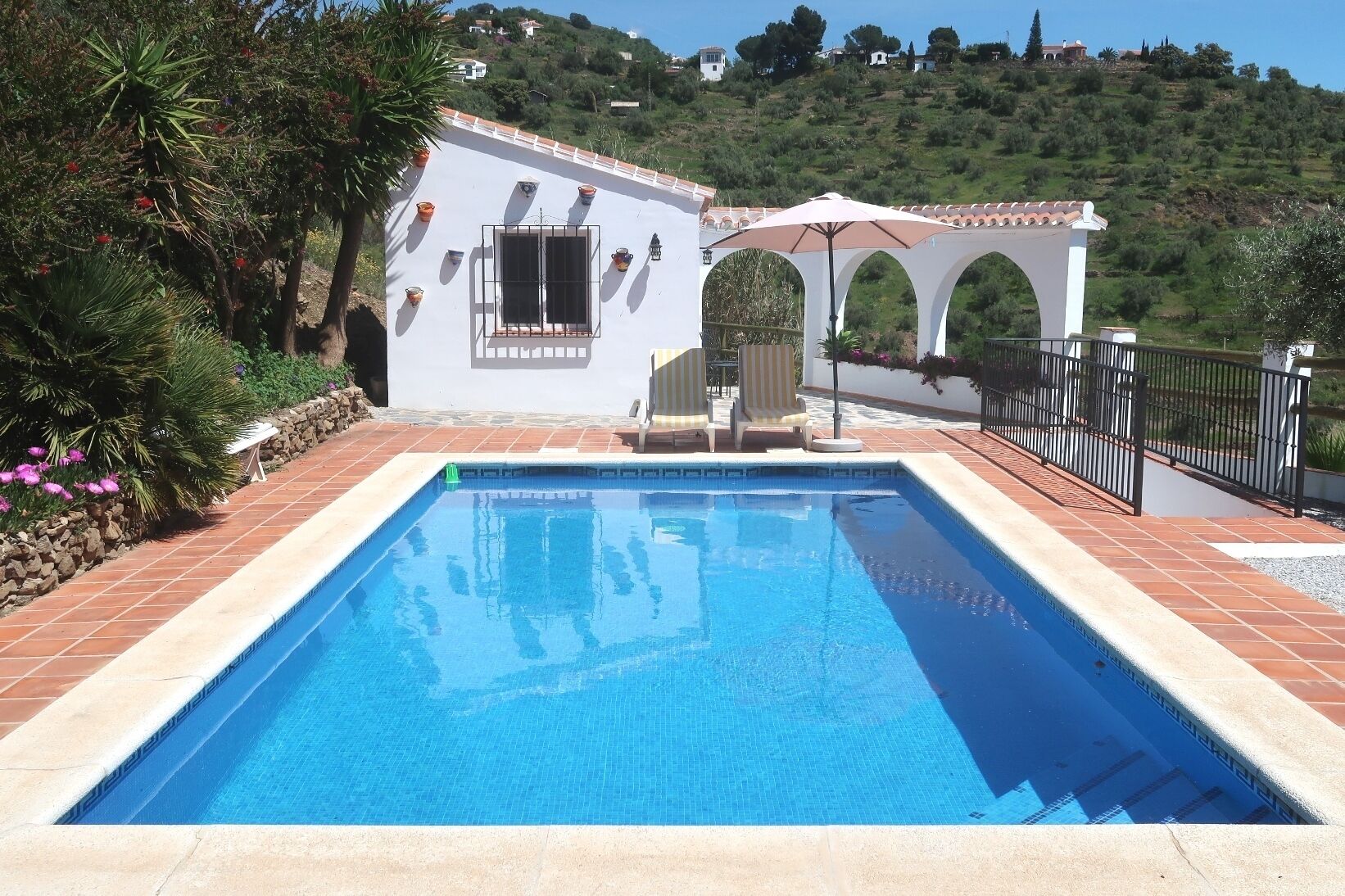 Une piscine rectangulaire bordée de carreaux, entourée d’un mur blanc percé de fenêtres et doté d’une porte. On y trouve un petit espace patio avec une table et des chaises, ainsi qu’une pergola avec un parasol.
