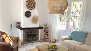 A living room with a fireplace, a sofa, a coffee table, and a wicker chair. - Villa Lantana - Private pool and sea views (Sayalonga)