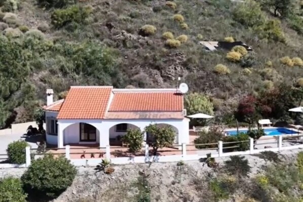 A white building with a red-tiled roof, a swimming pool, and a mountainous backdrop.