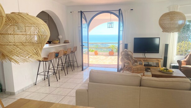 A modern living room with a large sofa, a dining area with wicker chairs, a TV, and a view of the ocean through an open archway. - Villa Lantana - Private pool and sea views (Sayalonga)