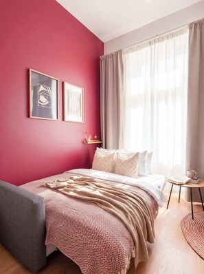 Une chambre à coucher comprenant un lit, une table de chevet, une chaise, une fenêtre avec des rideaux et deux tableaux encadrés accrochés au mur.
