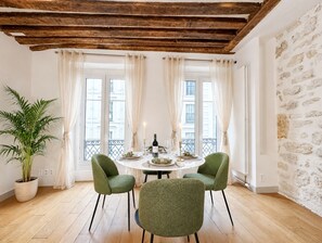 A dining area with a round table, two chairs, a potted plant, and a view of buildings through large windows. - 1550 Nazareth (Paris)