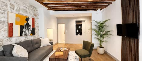 Un salon moderne avec un mur en pierre, un canapé gris, une table basse en bois, un fauteuil vert et un téléviseur à écran plat fixé sur un panneau en bois.