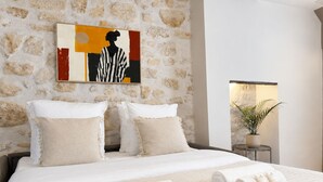 A hotel room with a stone wall, a bed with white bedding, two pillows, a painting, a bedside table with a plant, and rolled towels. - 1550 Nazareth (Paris)