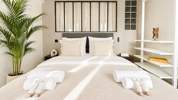 A hotel room with a large bed, a potted plant, a nightstand, and a shelf with a Louis Vuitton box. - 1550 Nazareth (Paris)