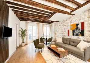 A modern living room with exposed wooden beams, a flat-screen TV, a sofa, a coffee table, and a dining area with chairs and a table. - 1550 Nazareth (Paris)