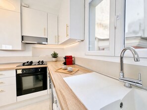 A modern kitchen with white cabinets, a stainless steel range hood, a built-in oven, a coffee maker, and a window above the sink. - 1550 Nazareth (Paris)