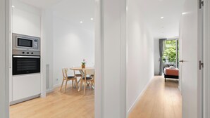 A modern kitchen with built-in appliances, a dining area with a round table and chairs, and a hallway leading to a bedroom with a view of greenery.