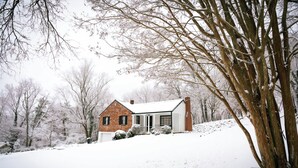 A snow-covered house with a brick exterior and a white roof, surrounded by bare trees and a snowy landscape. - Stay Roanoke | Cute 3BR Near Blue Ridge Parkway (Roanoke)