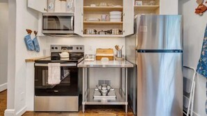 A compact kitchen with a microwave, oven, and stainless steel refrigerator. There are open cabinets above the counter, and a small table with kitchen utensils. - Stay Roanoke | Cute 3BR Near Blue Ridge Parkway (Roanoke)