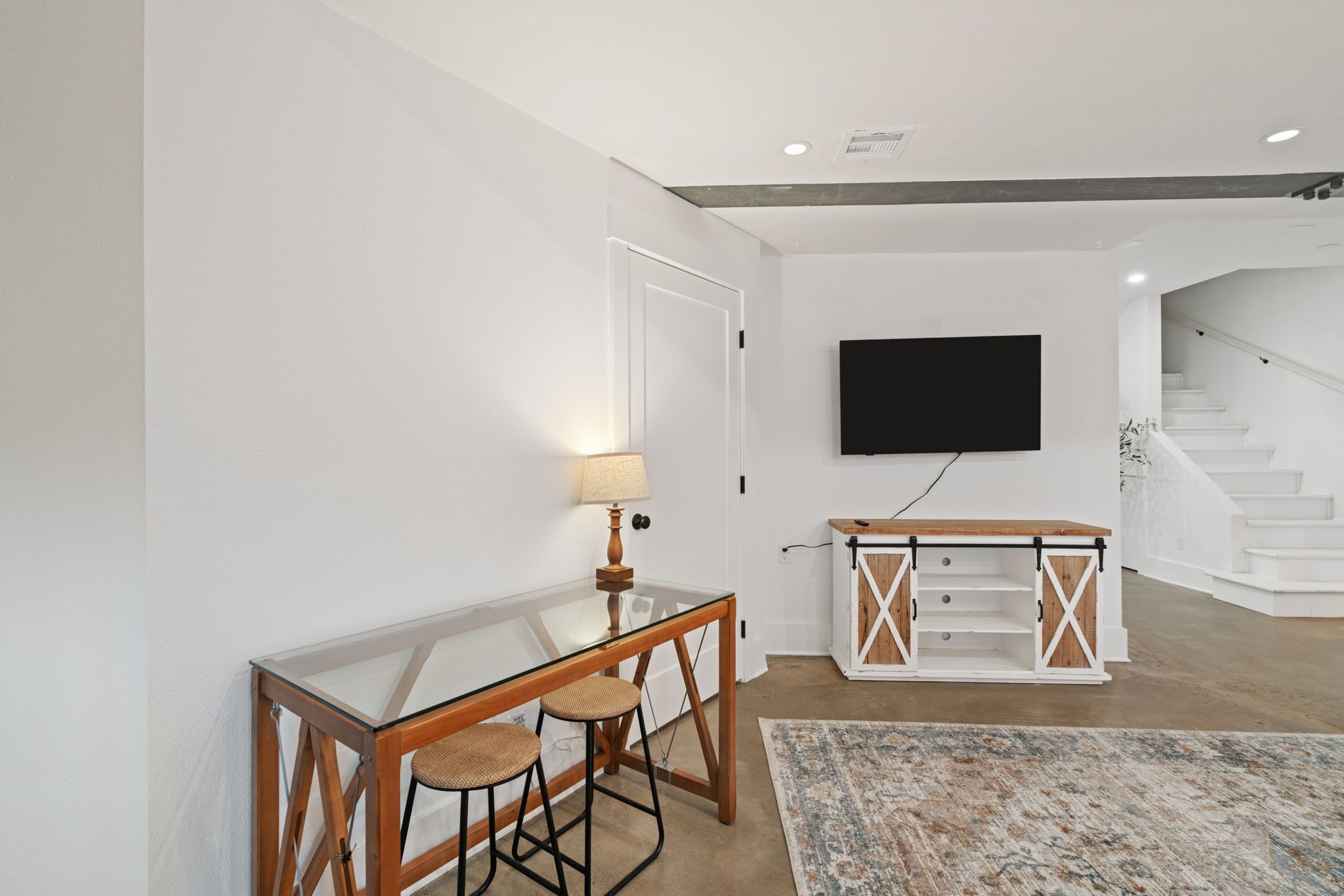 A modern living room with a glass coffee table, two bar stools, a mounted TV, and a staircase leading to another level.