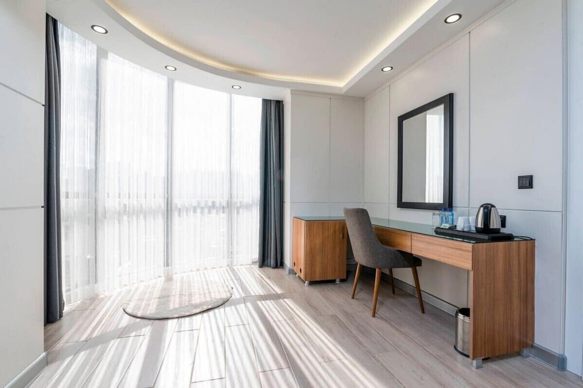 A modern hotel room with a wooden desk, a chair, a large mirror, and a view through floor-to-ceiling windows.