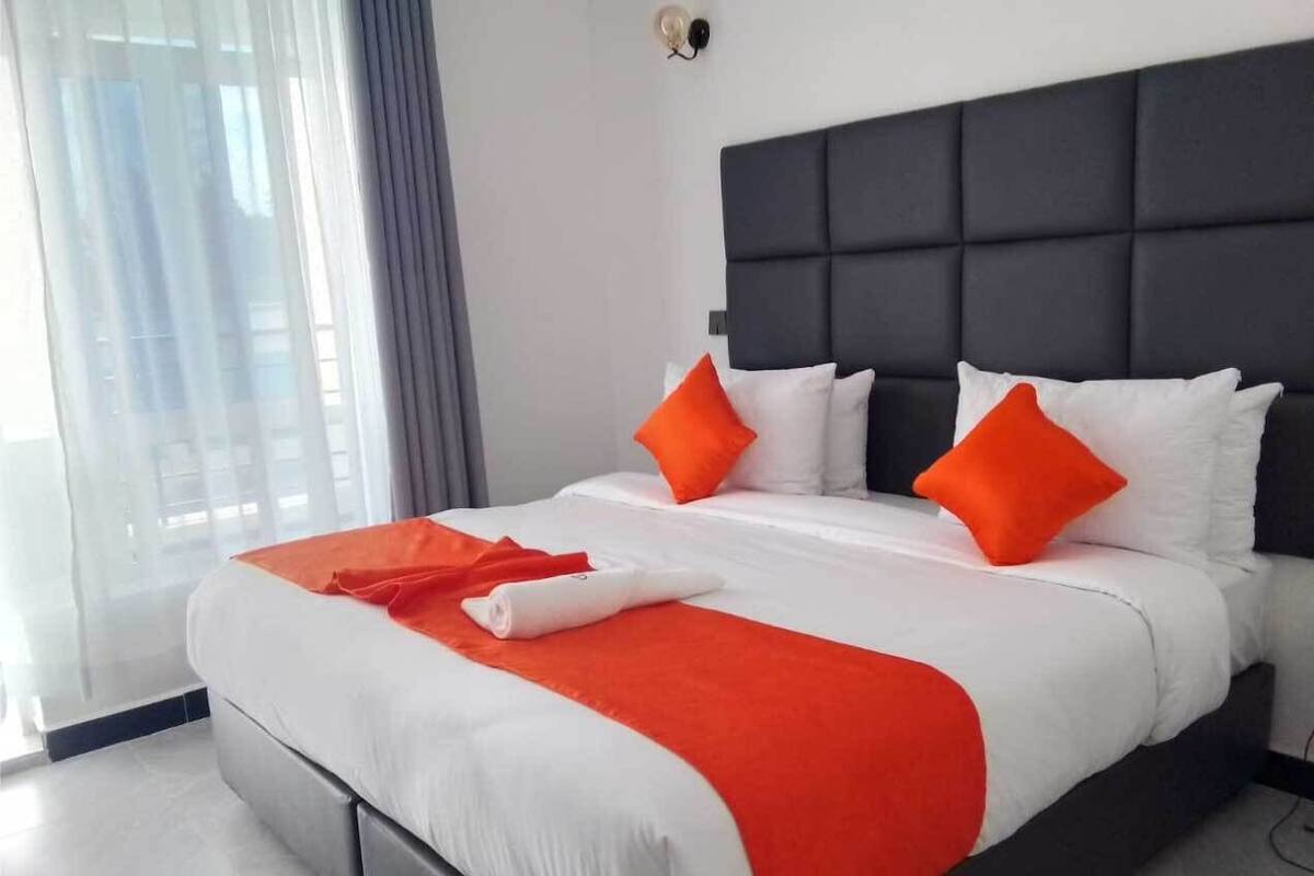 A modern hotel room with a large bed, white bedding, and orange accents.